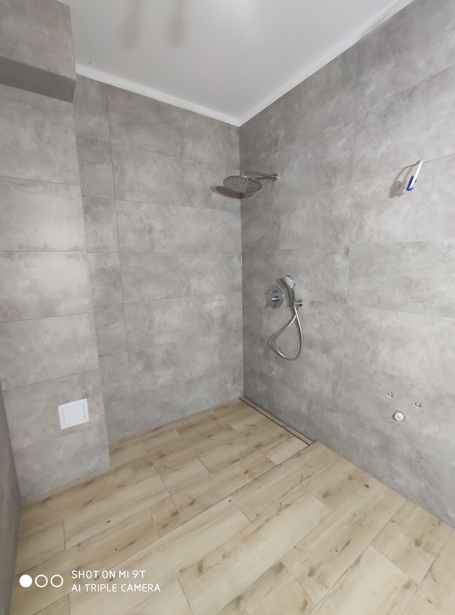 Nowoczesna kabina prysznicowa z szarymi płytkami imitującymi beton i drewnopodobną podłogą, widoczny odpływ liniowy, deszczownica i bateria termostatyczna, zasilanie do hydromasażu.
