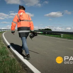 Mężczyzna w odblaskowej kurtce idzie poboczem drogi z aparaturą pomiarową na tle błękitnego nieba z chmurami.