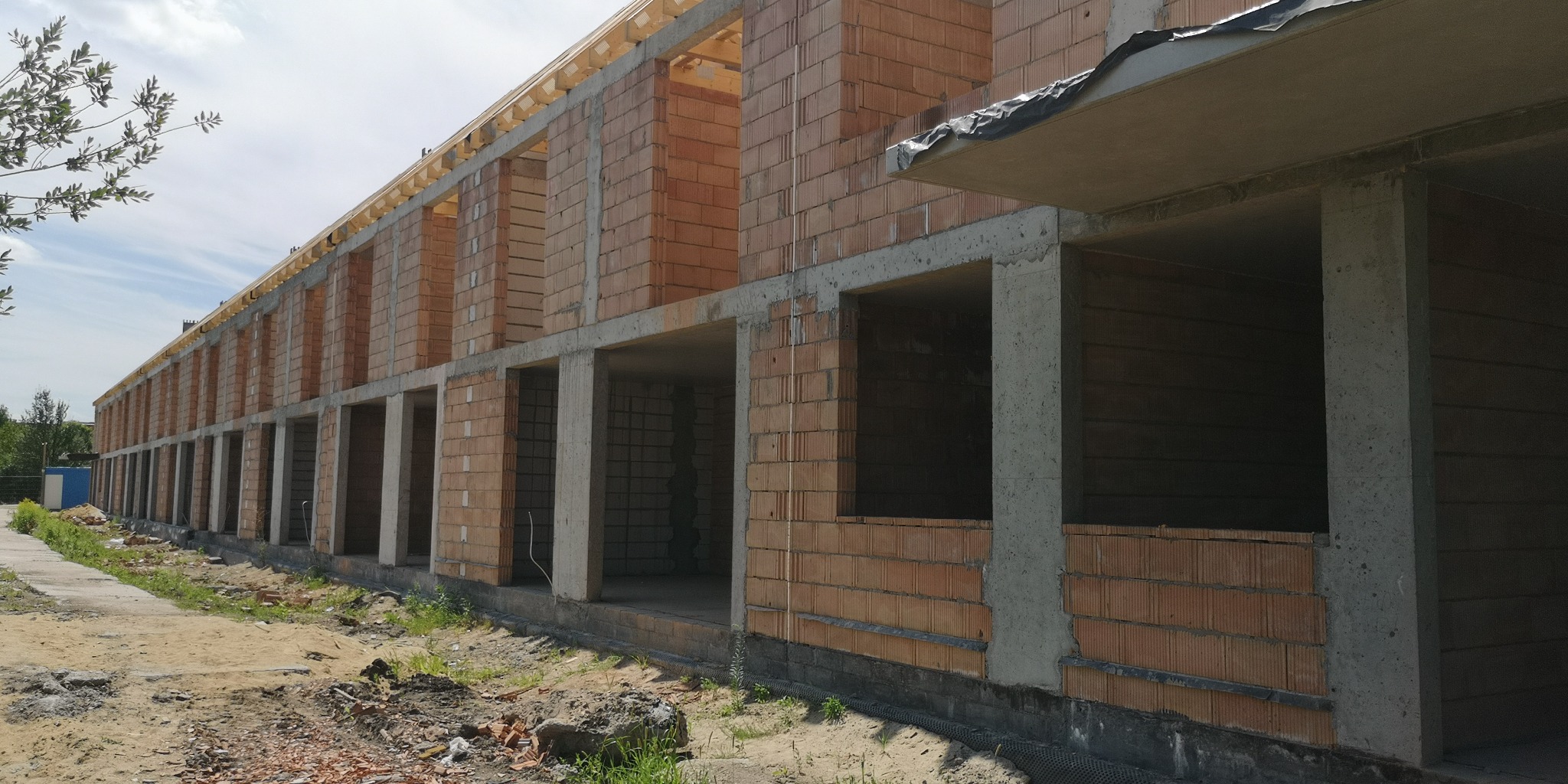 Wznoszenie budynku wielorodzinnego z czerwonej cegły. Widoczne surowe ściany i betonowe filary konstrukcyjne. Budowa w toku, zarys przyszłych okien i balkonów.