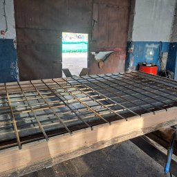 ARDSWELD Arkadiusz Dopierała - Klatka zbrojeniowa z prętów stalowych, przygotowana na drewnianym stole w warsztacie, widoczny otwarty garaż z zielonym kontenerem na zewnątrz.