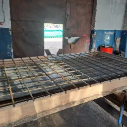 Klatka zbrojeniowa z prętów stalowych, przygotowana na drewnianym stole w warsztacie, widoczny otwarty garaż z zielonym kontenerem na zewnątrz.