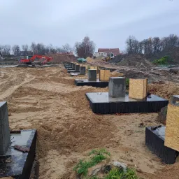 Przygotowane betonowe fundamenty pod budynek z deskowaniem, widoczny ciężki sprzęt budowlany i usypane pryzmy ziemi w tle, pochmurny dzień.