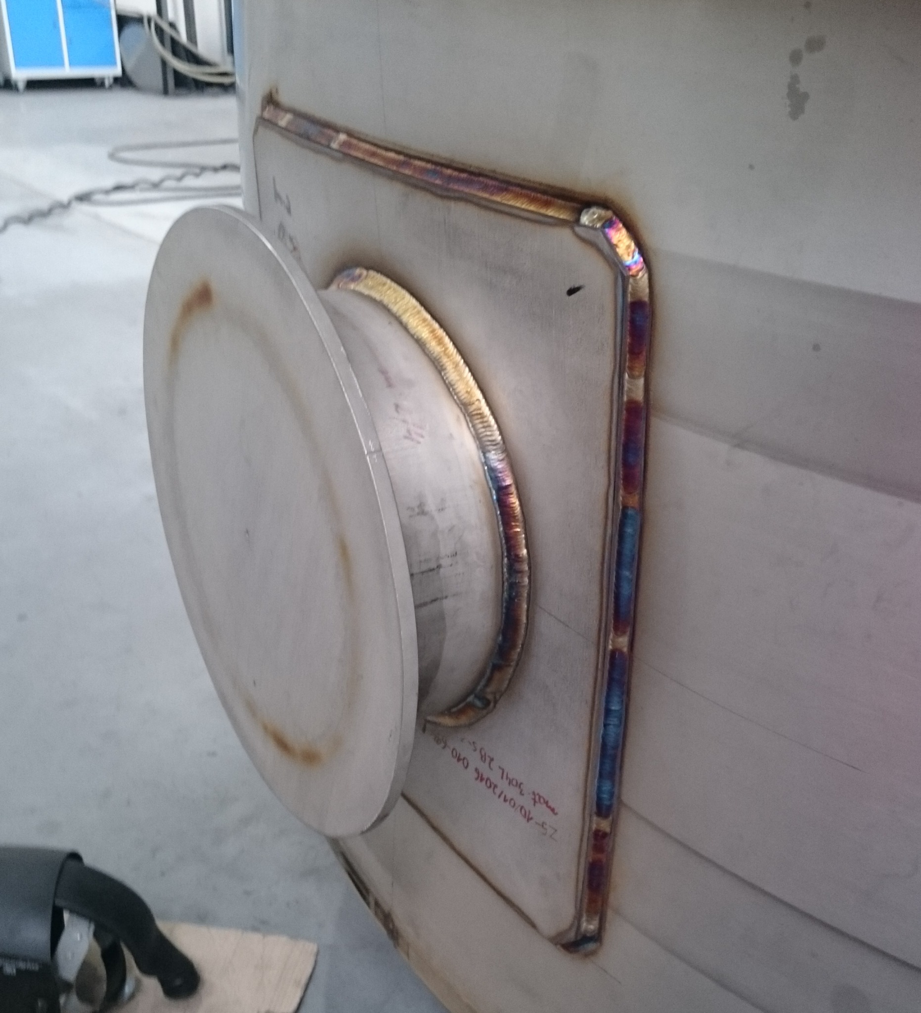 Metalowa konstrukcja z okrągłym elementem przymocowanym do kwadratowej płyty, widoczne spawy z opalizującymi przebarwieniami, notatki odręczne na powierzchni.