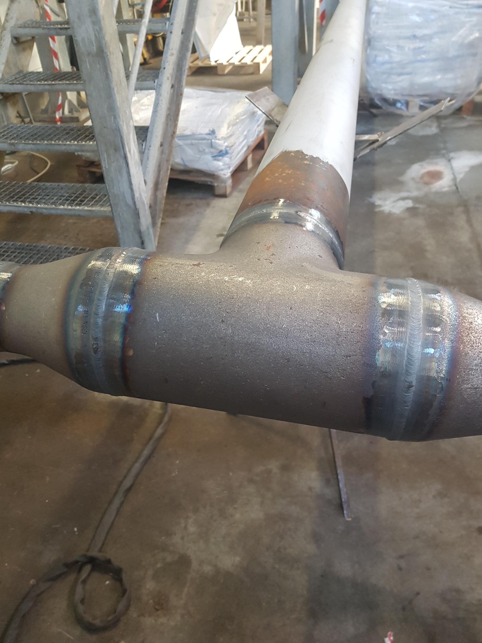 Spojenie spawane rur stalowych w kształcie litery T z widocznymi śladami spawania i oznaczeniami na powierzchni, w tle schody metalowe i materiały budowlane.