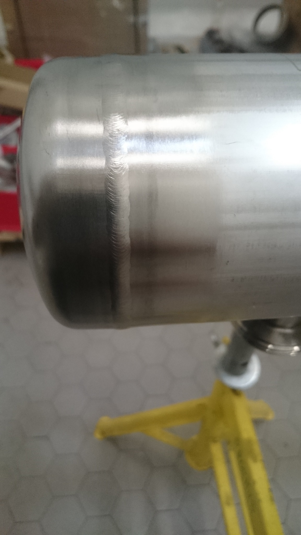 Zbliżenie na spawany szew na metalowym zbiorniku, widoczny precyzyjny ślad spawania TIG na gładkiej, srebrzystej powierzchni, zbiornik umieszczony na żółtym stojaku.