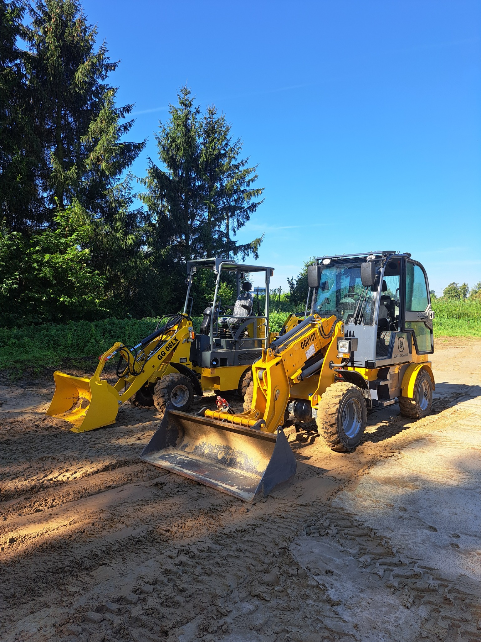 Dwa żółte miniładowarki na nierównym, ubitym terenie, prawdopodobnie po niwelacji, z widocznymi śladami opon i łyżek; w tle wysokie drzewa iglaste i błękitne niebo.