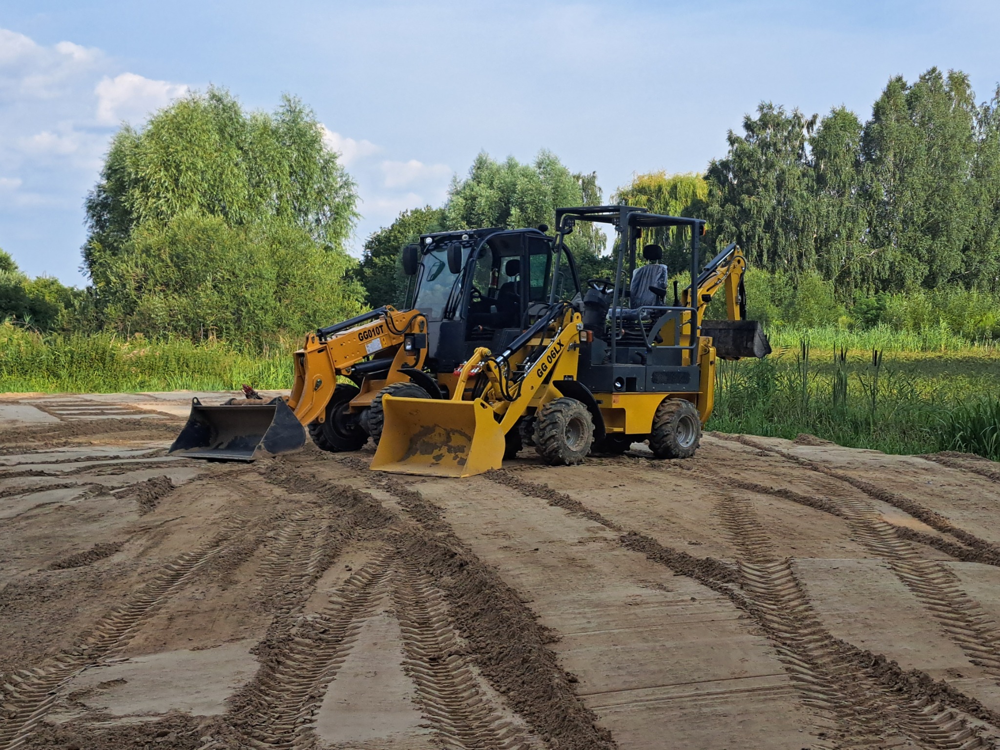Dwie żółte miniładowarki GG010T i GG 06LX na wyrównanym, piaszczystym terenie z widocznymi śladami opon, w tle zielona roślinność i drzewa pod błękitnym niebem.
