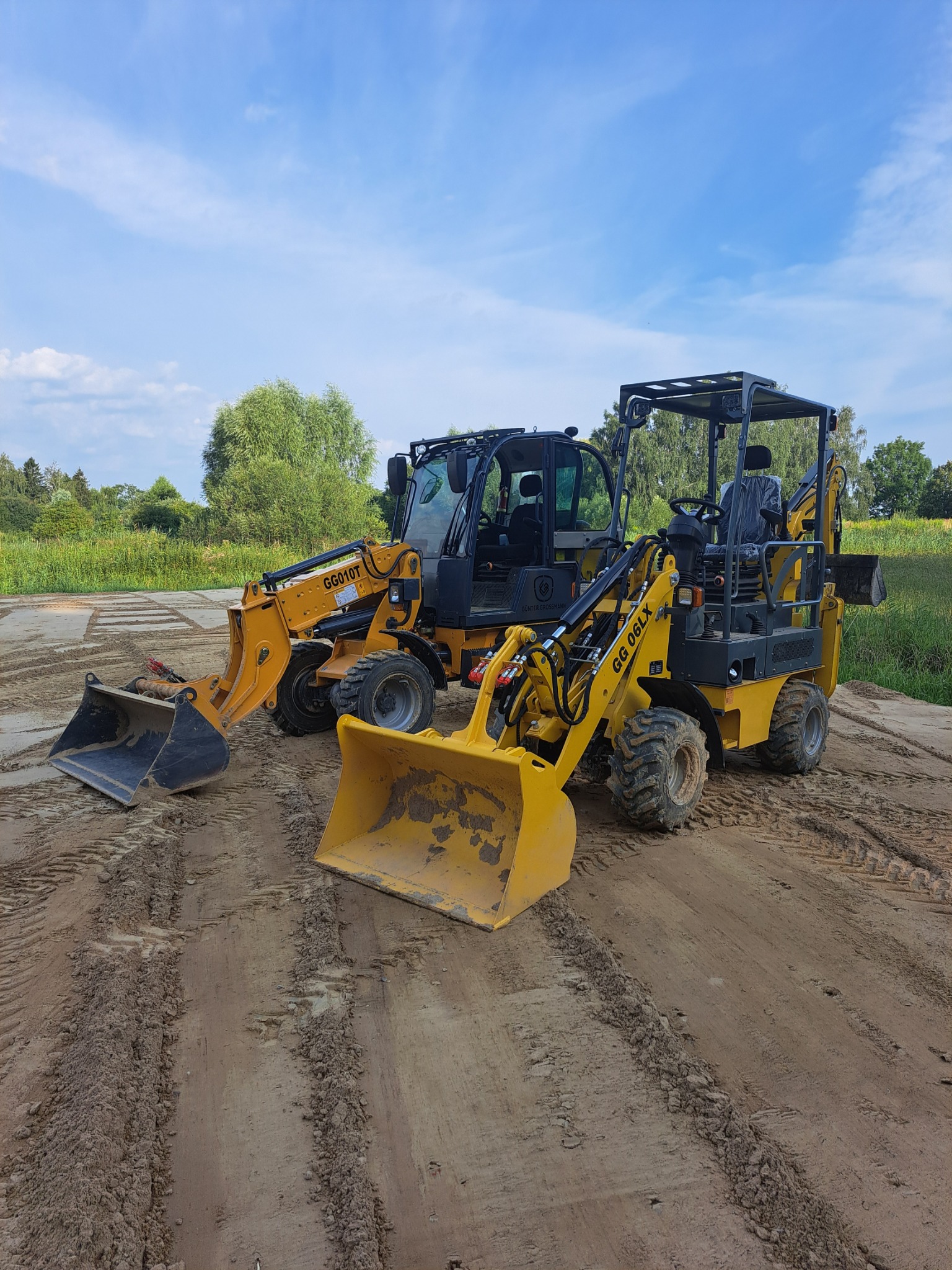 Dwa żółte miniładowarki Gunter Grossmann GG010T i GG 06LX stoją na ubitej ziemi z widocznymi śladami opon. Jeden z nich ma otwartą kabinę, a drugi zamkniętą. W tle widać drzewa i błękitne niebo...