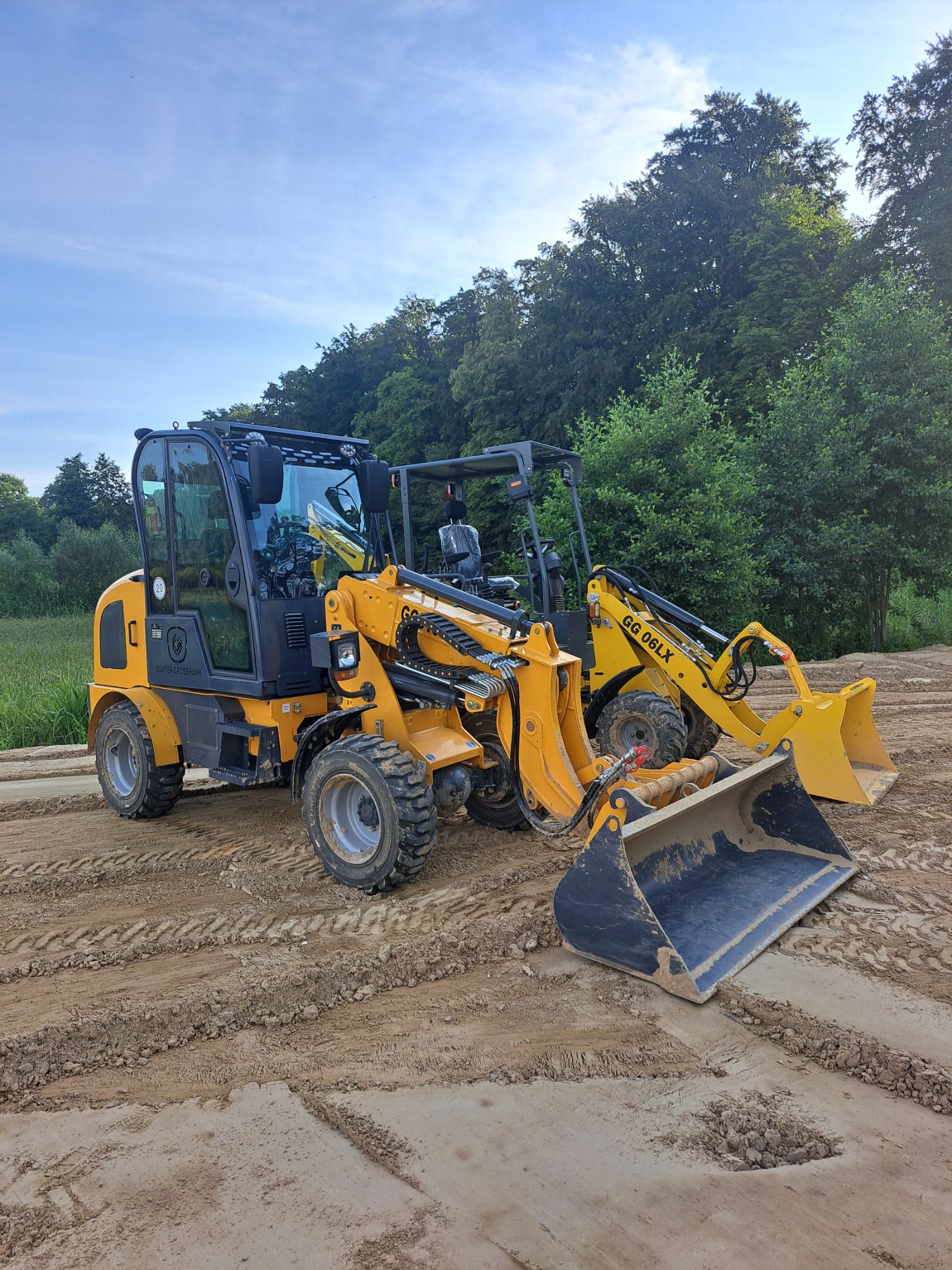 Dwa żółte miniładowarki Gunter Grossmann na ubitym, piaszczystym terenie z widocznymi śladami opon, w tle zielone drzewa i błękitne niebo.