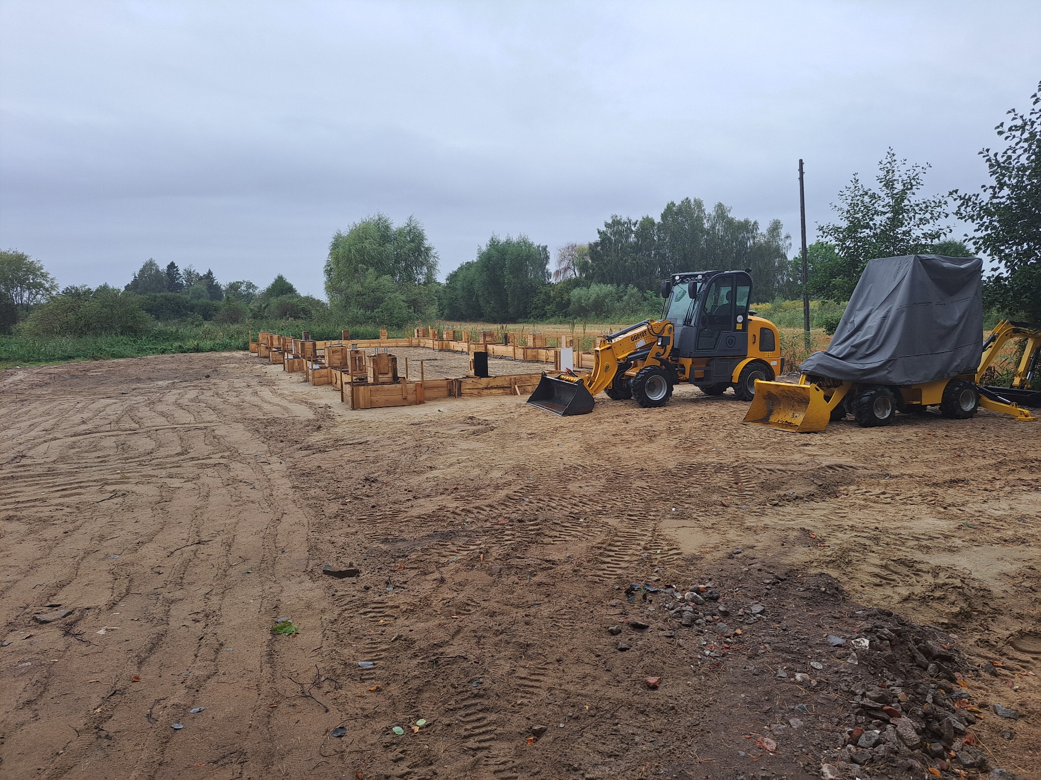 Przygotowany teren pod fundamenty z drewnianym szalunkiem i żółtą mini ładowarką na ziemnym placu budowy w pochmurny dzień.
