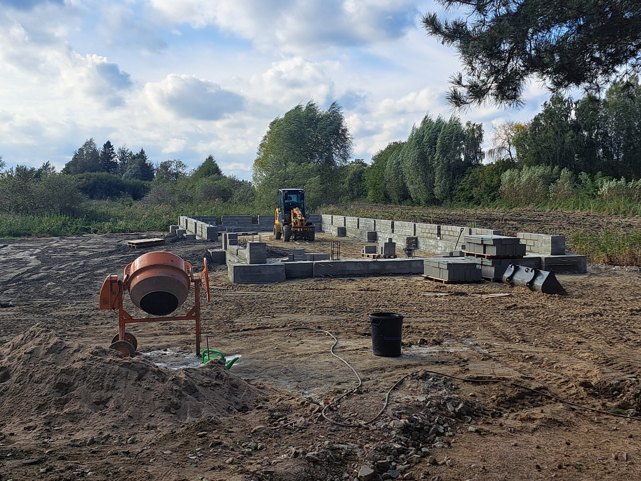 Wykop pod fundamenty z widoczną częścią zbrojenia, betoniarka na pierwszym planie i mała koparka w tle, teren przygotowany pod budowę, pochmurne niebo.