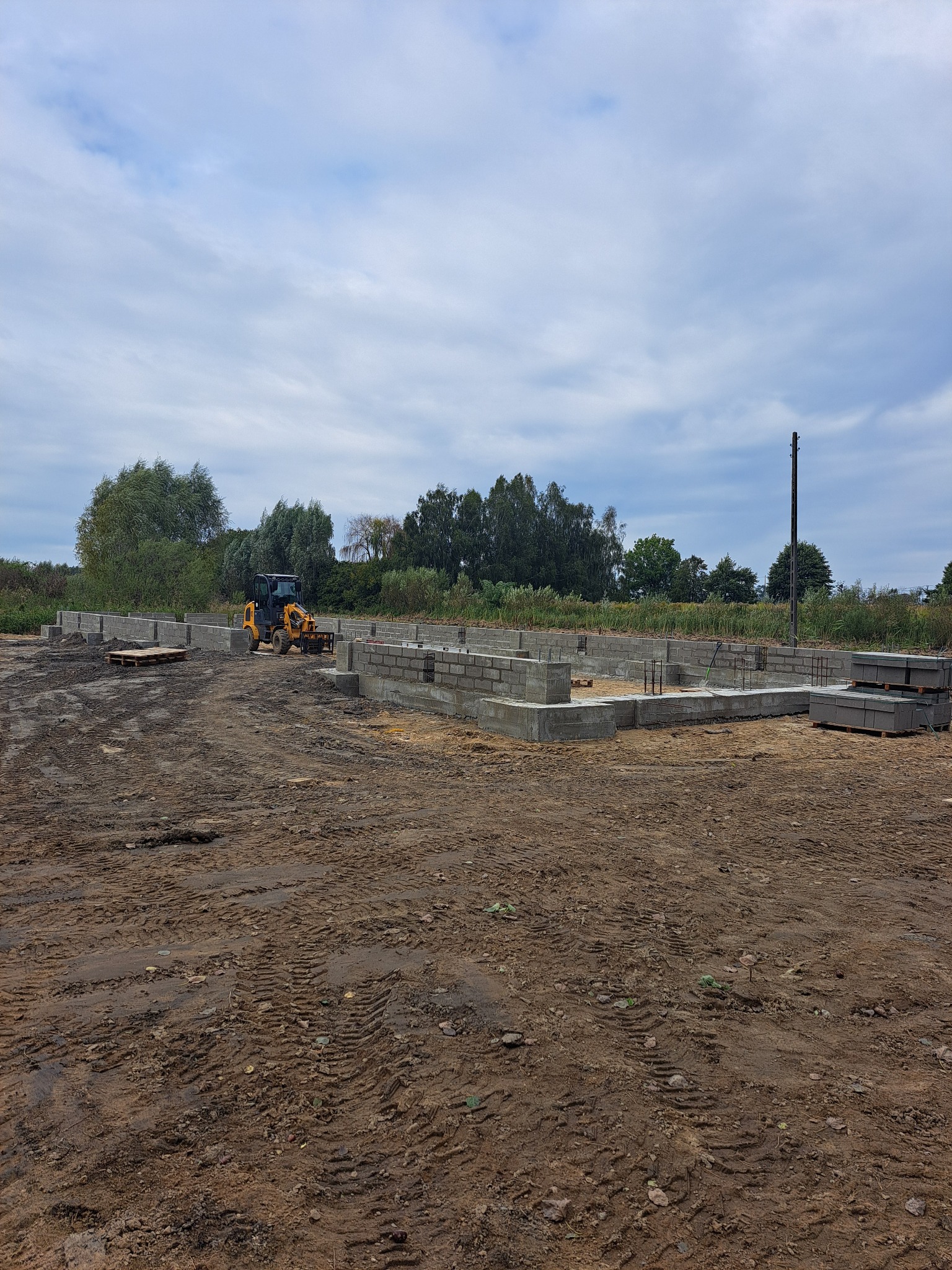 Żółta ładowarka teleskopowa na placu budowy, fundamenty z bloczków betonowych częściowo wylane, widoczne zbrojenia, teren wyrównany, ślady opon na ziemi, pochmurne niebo.