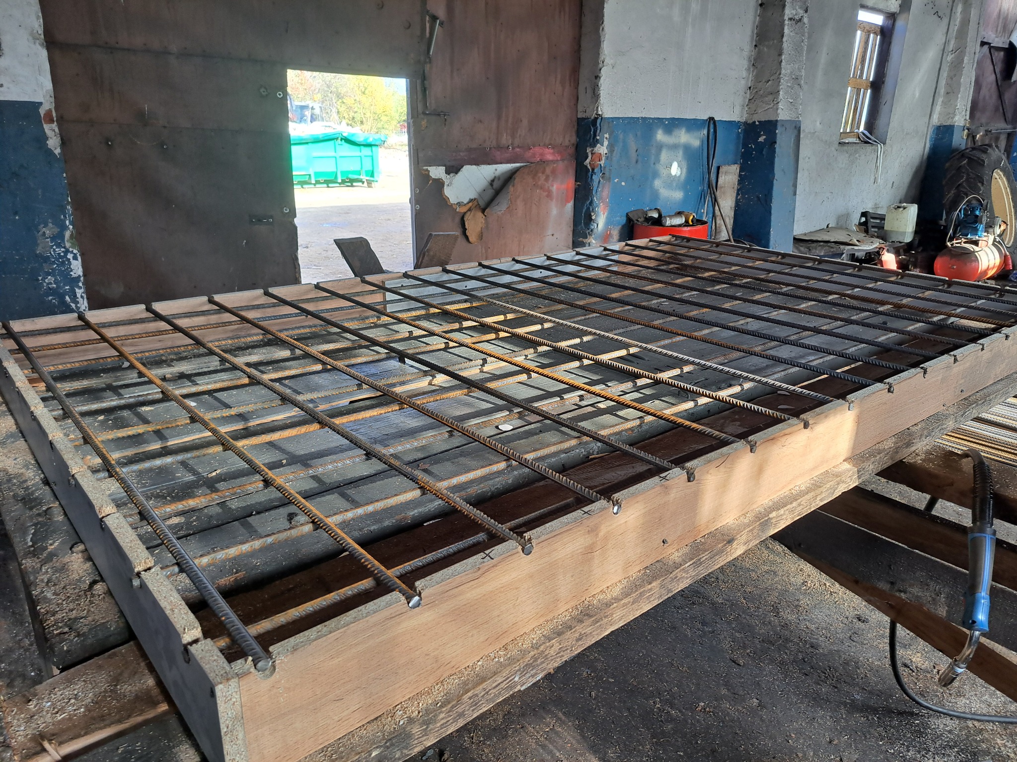 Konstrukcja z prętów zbrojeniowych, tworząca siatkę, umieszczona na drewnianej ramie w warsztacie, widoczne ślady spawania i narzędzia.