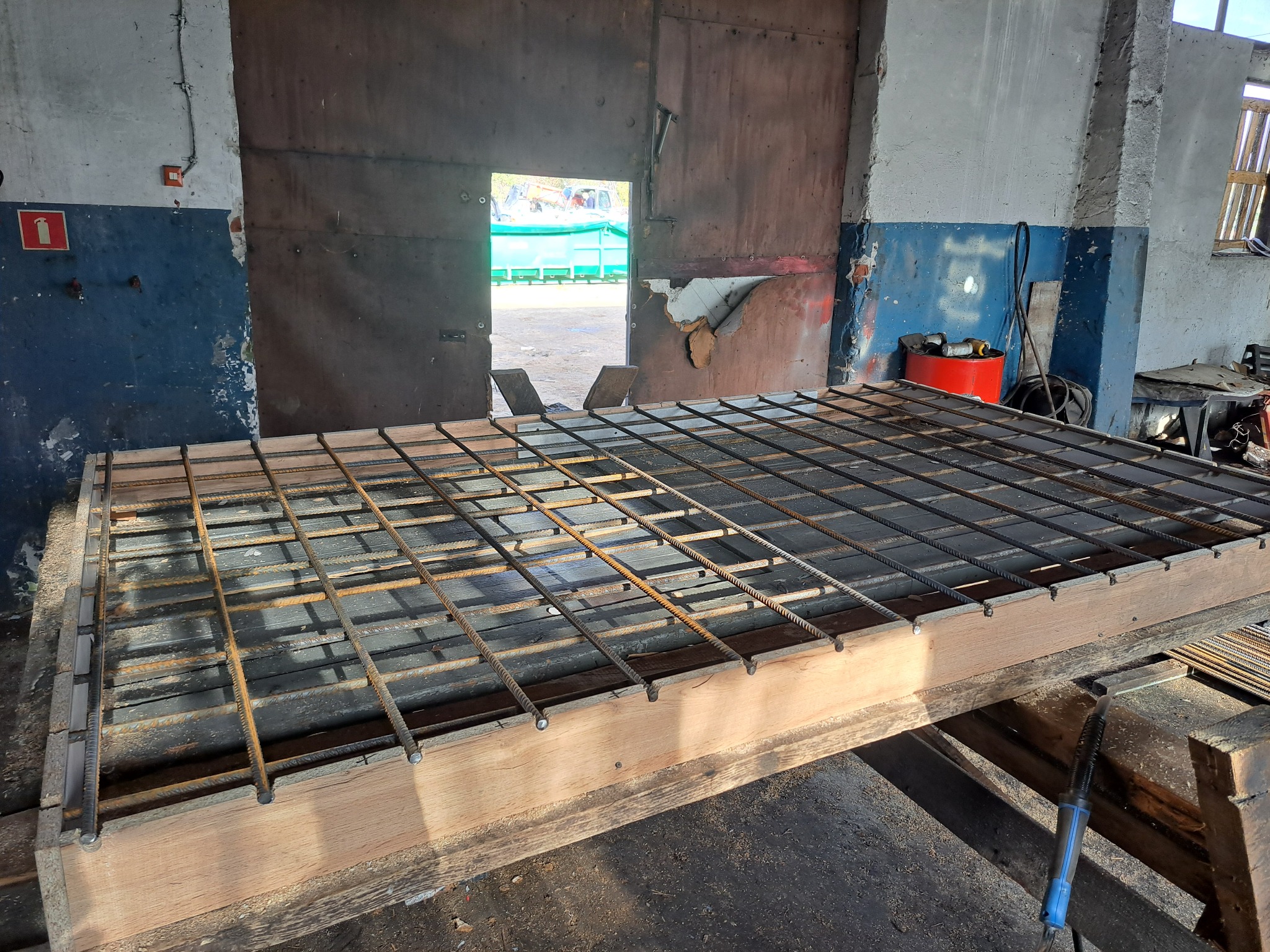 Klatka zbrojeniowa z prętów stalowych, przygotowana na drewnianym stole w warsztacie, widoczny otwarty garaż z zielonym kontenerem na zewnątrz.