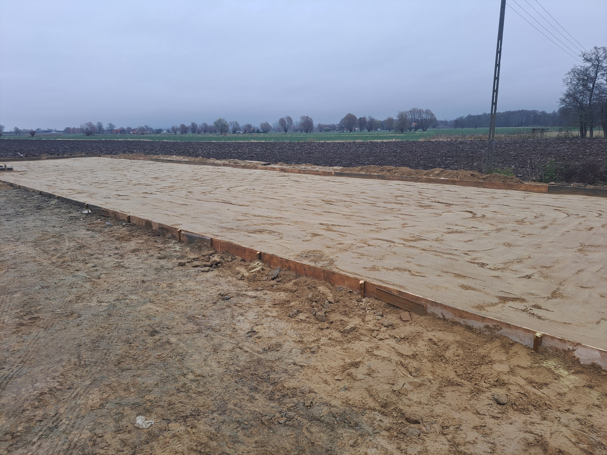 Przygotowany teren pod budowę z drewnianym obramowaniem, widok na pole i pochmurne niebo w tle.