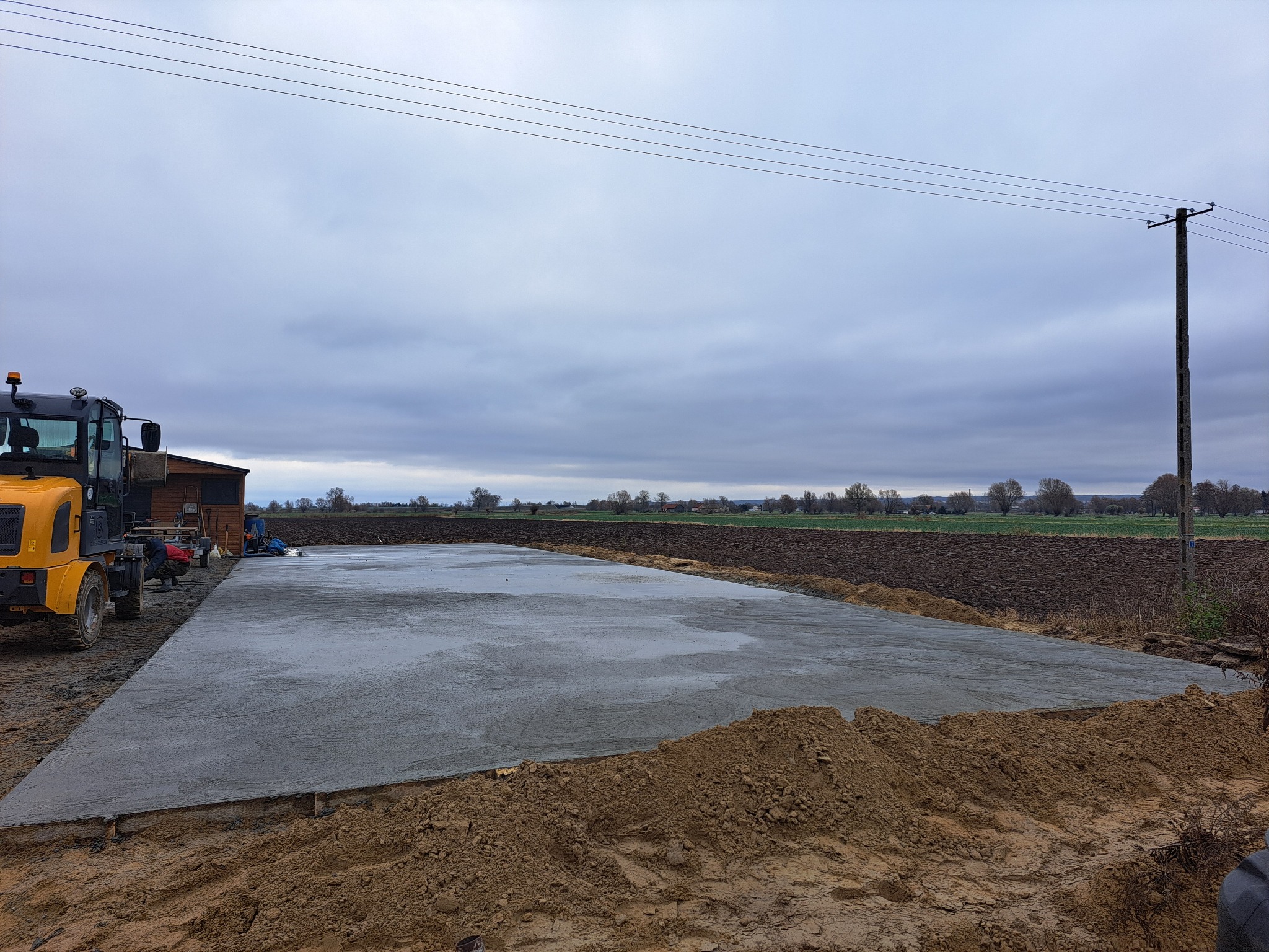 Wylany, świeży betonowy fundament pod budowę hali, widoczny częściowo żółty sprzęt budowlany i pracownicy, w tle pole i pochmurne niebo.