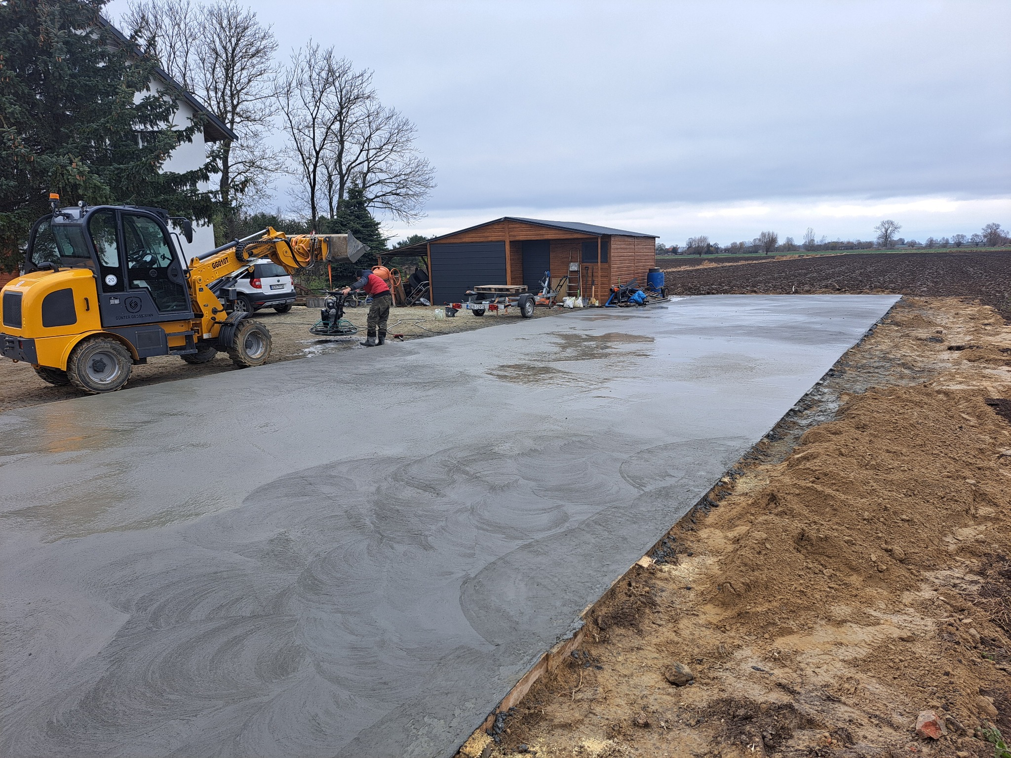 Świeżo wylana, gładka betonowa posadzka na zewnątrz, wyrównywana przez pracownika zacieraczką mechaniczną, obok żółta ładowarka teleskopowa, w tle budynek gospodarczy i pole uprawne pod pochmurnym...