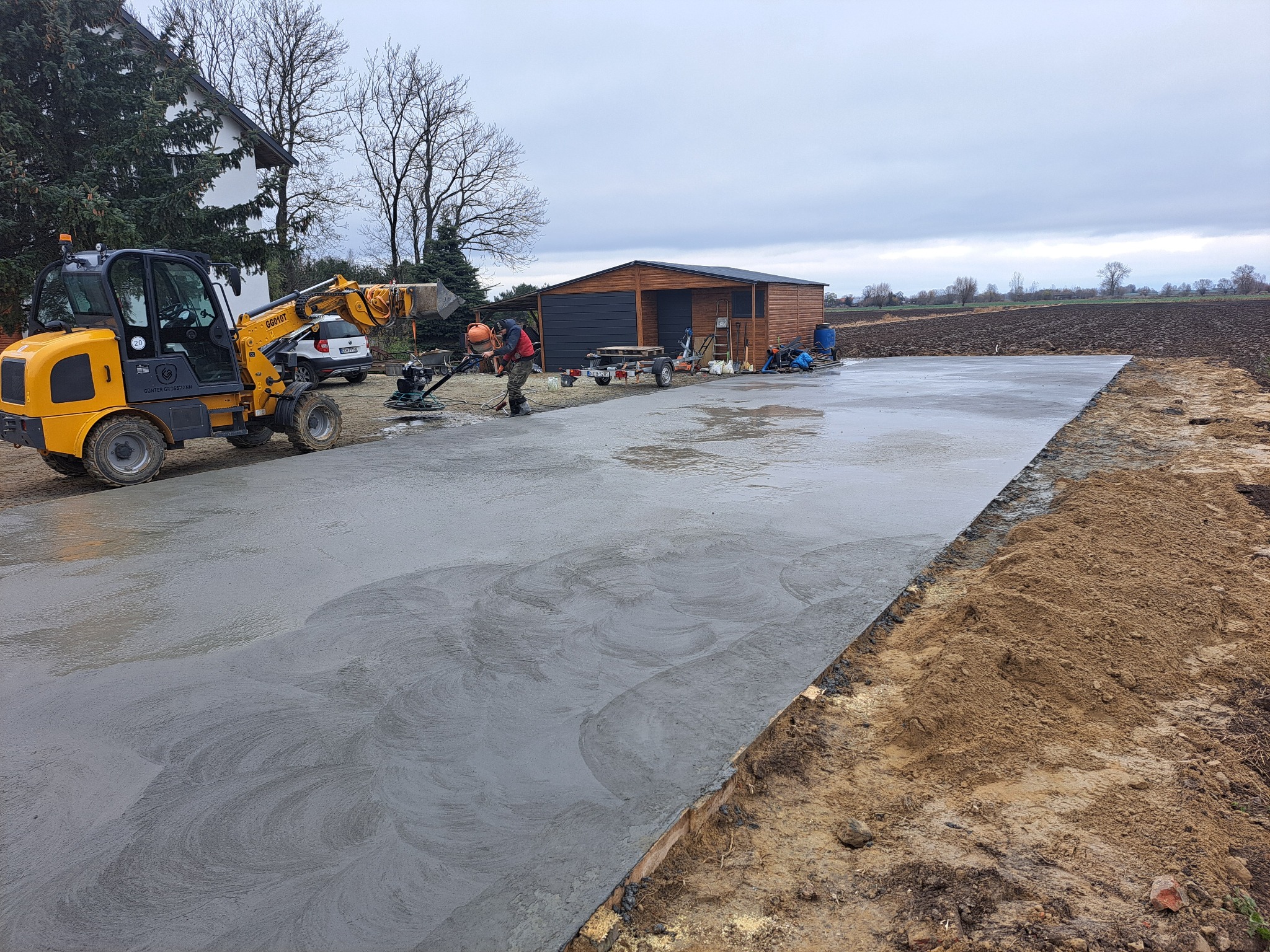 Świeżo wylana, gładka betonowa powierzchnia z widocznym wzorem obróbki, obok stoi żółta mini-ładowarka i pracownik zacierający beton, w tle budynek gospodarczy i pole.