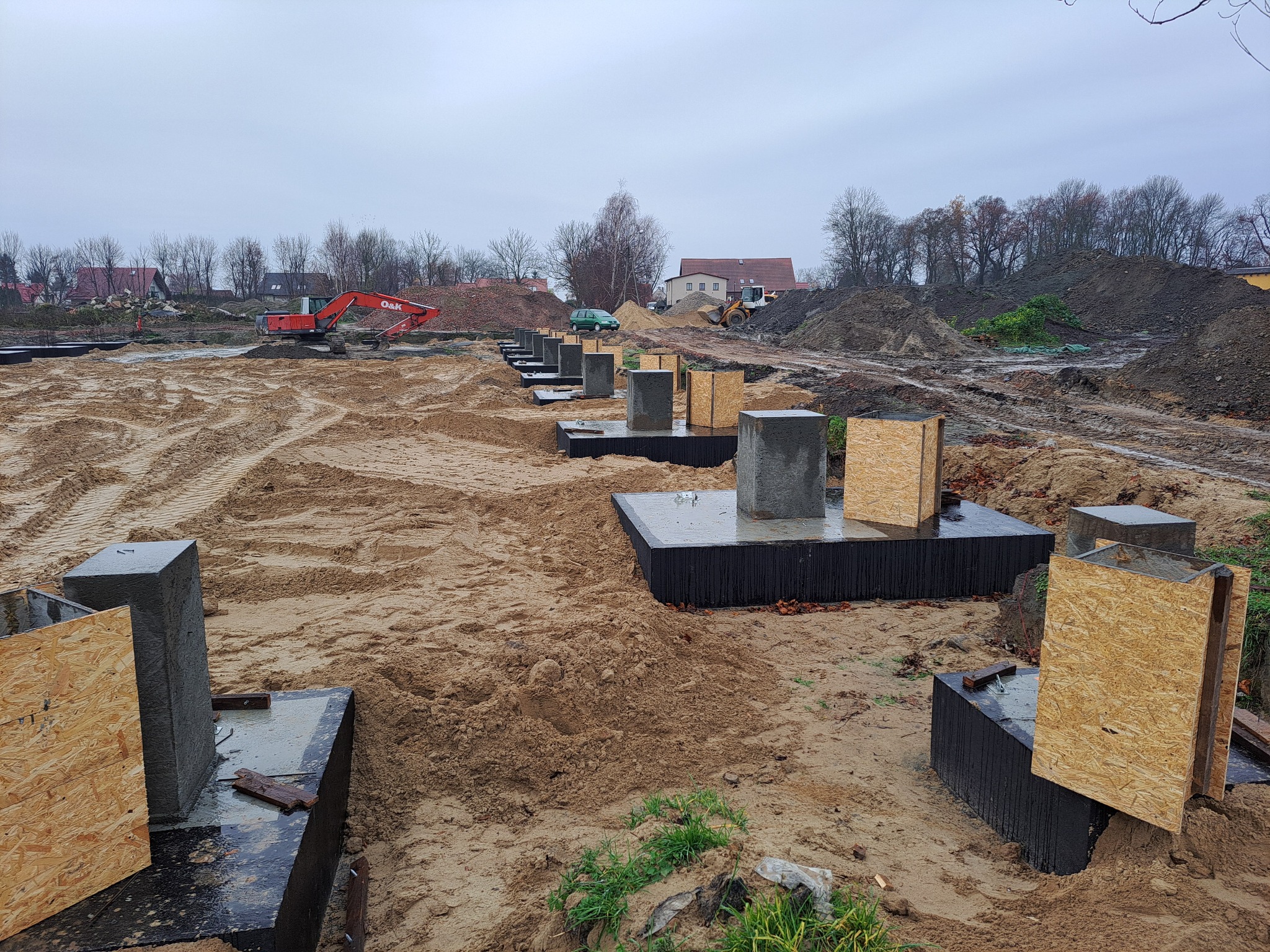 Przygotowane betonowe fundamenty pod budynek z deskowaniem, widoczny ciężki sprzęt budowlany i usypane pryzmy ziemi w tle, pochmurny dzień.