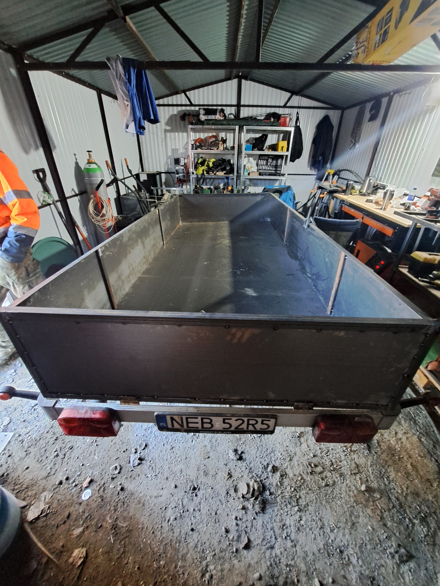 Spawana, metalowa przyczepa transportowa w warsztacie, widok z tyłu, z widoczną tablicą rejestracyjną i elementami konstrukcyjnymi spawanymi punktowo.