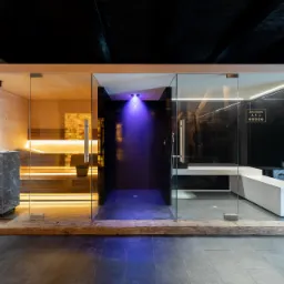 Nowoczesna sauna z trzema strefami: drewniana sauna z kamiennym piecem i oświetleniem, ciemna strefa relaksu z niebieskim światłem oraz szklana łaźnia parowa z białą ławką.