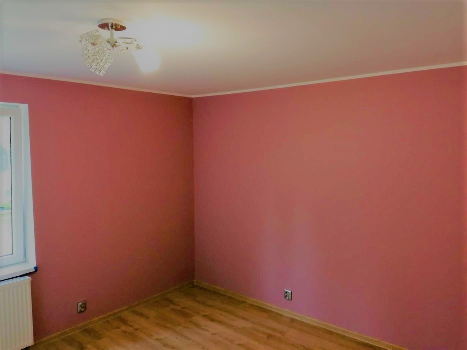 Pomalowany pokój na kolor brudny róż, z drewnianą podłogą, białym sufitem i ozdobną lampą sufitową.