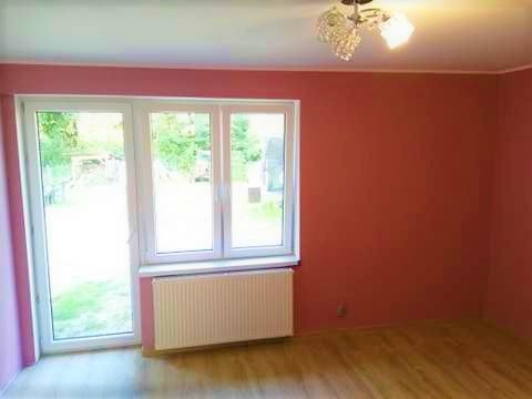 Pusty pokój z pomarańczowymi ścianami, białym oknem i grzejnikiem pod nim oraz drewnianą podłogą, oświetlony lampą sufitową z kryształkami.