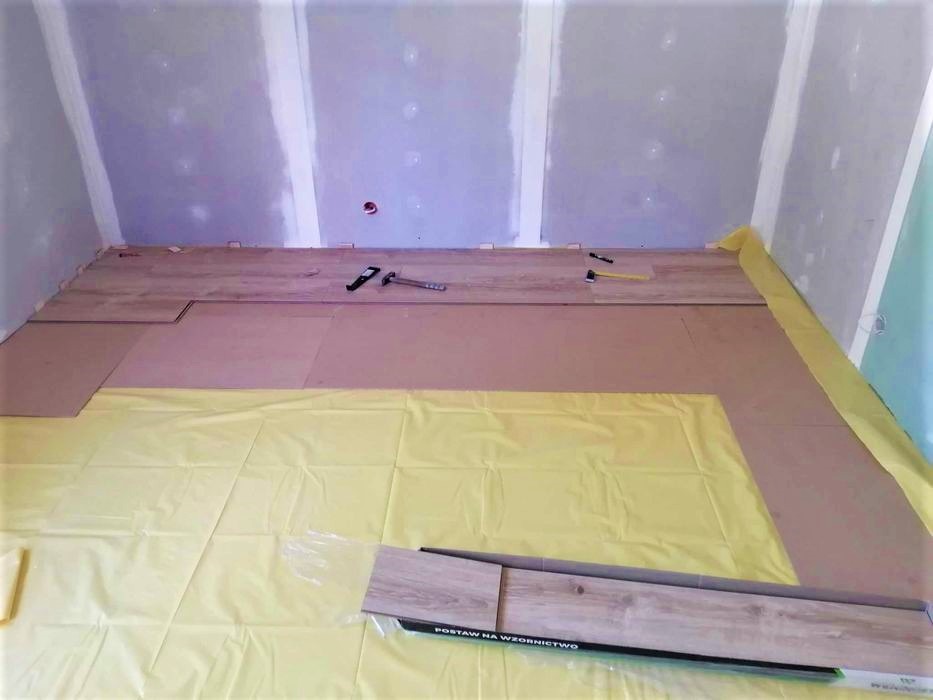 Układanie paneli podłogowych w pomieszczeniu z przygotowanymi ścianami i podłożem zabezpieczonym folią, widoczne narzędzia i opakowanie paneli.