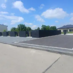 Nowoczesne, czarne ogrodzenie panelowe z poziomych desek, oddzielające posesję z domem jednorodzinnym od ulicy; brama wjazdowa z szarych paneli. Kostka brukowa w dwóch odcieniach szarości.