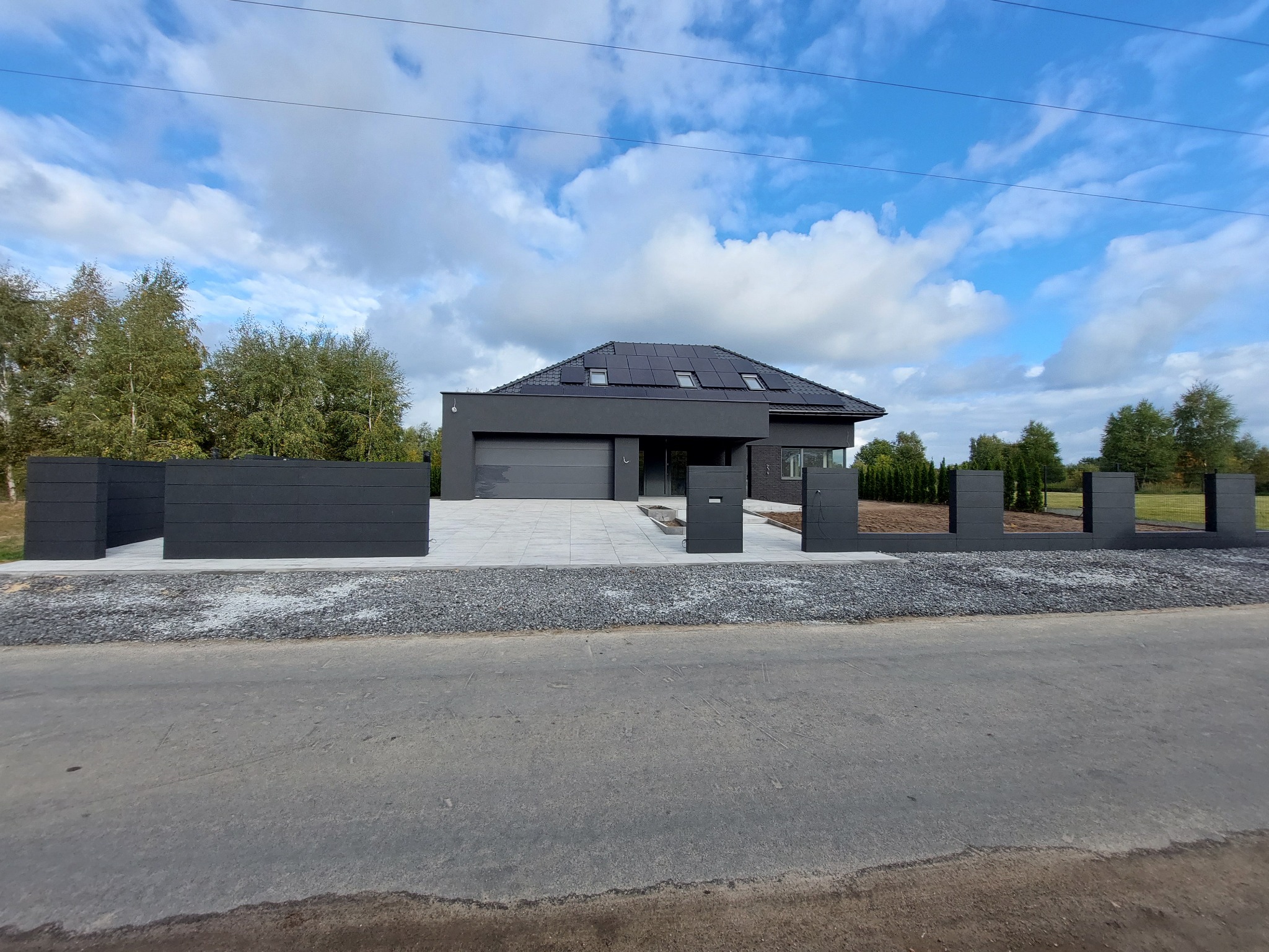 Nowoczesny dom z płaskim dachem i panelami słonecznymi, otoczony grafitowym ogrodzeniem modułowym z betonową podmurówką i kostką brukową, widok z ulicy w słoneczny dzień.