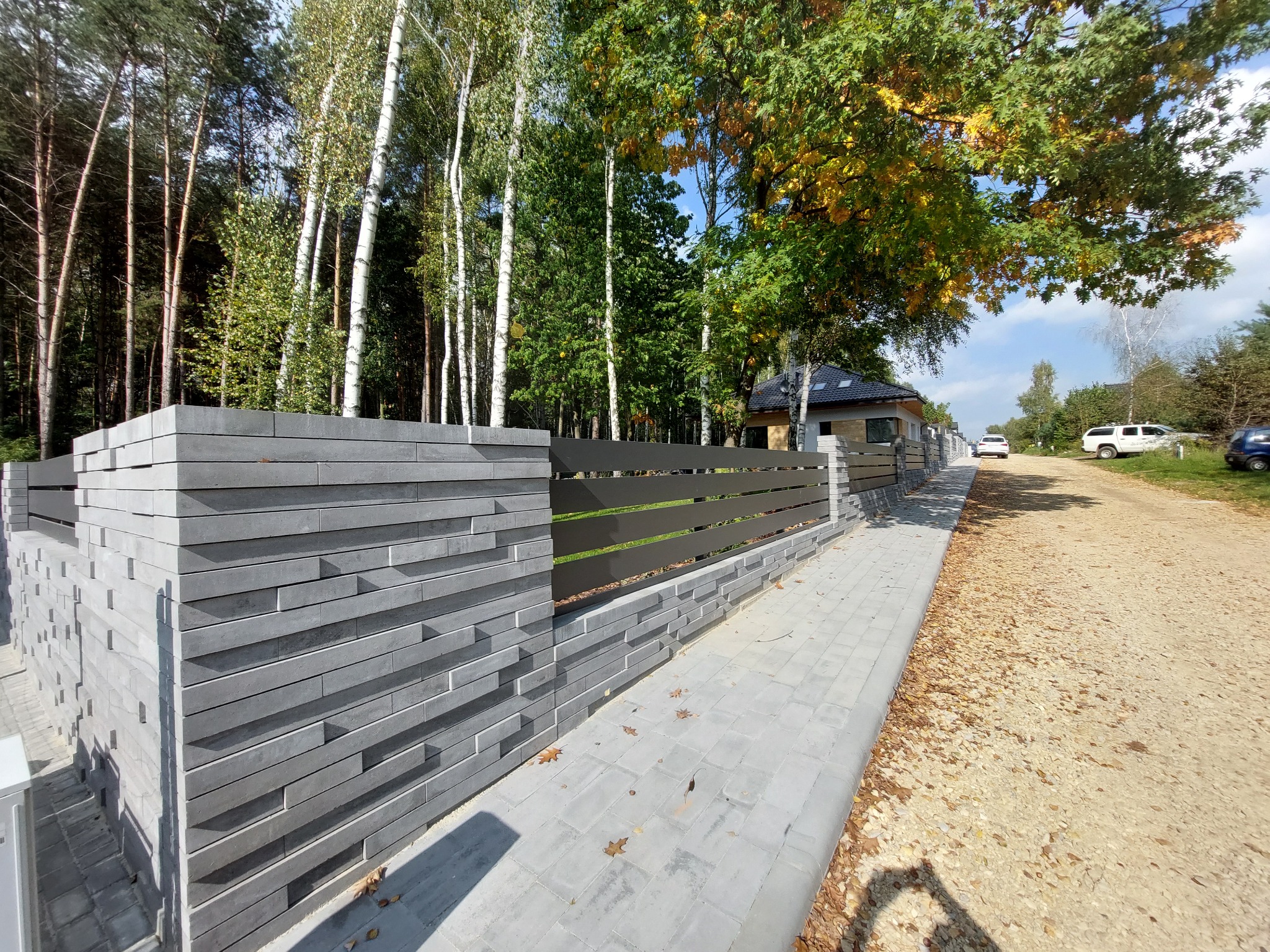 Nowoczesne ogrodzenie z szarej kostki betonowej z poziomymi metalowymi przęsłami, widok z perspektywy chodnika, w tle drzewa i dom jednorodzinny.