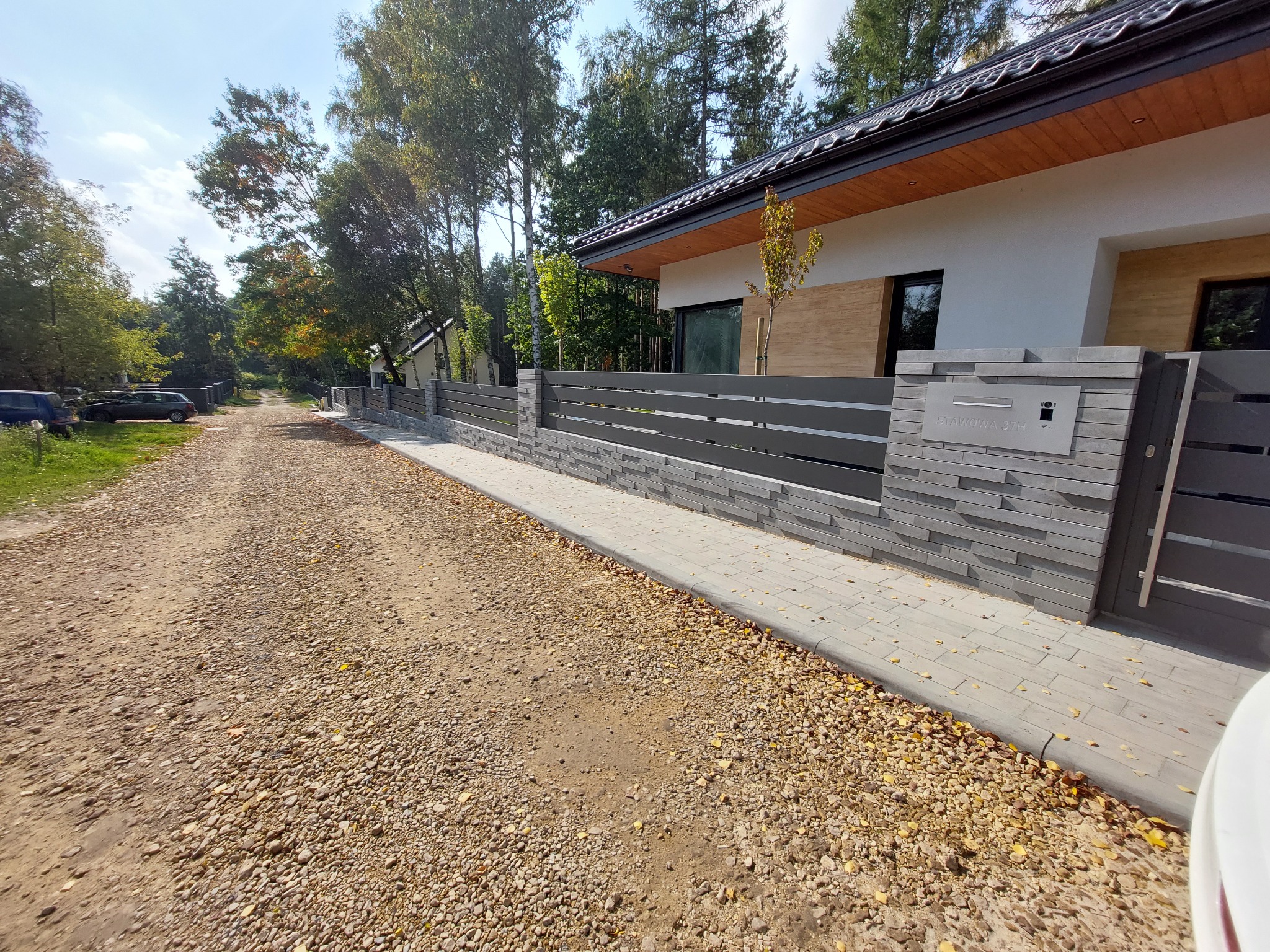 Nowoczesne ogrodzenie panelowe z szarej cegły, zintegrowane ze skrzynką pocztową o minimalistycznym designie, na tle domu z elementami drewnianej elewacji i żwirowej drogi.