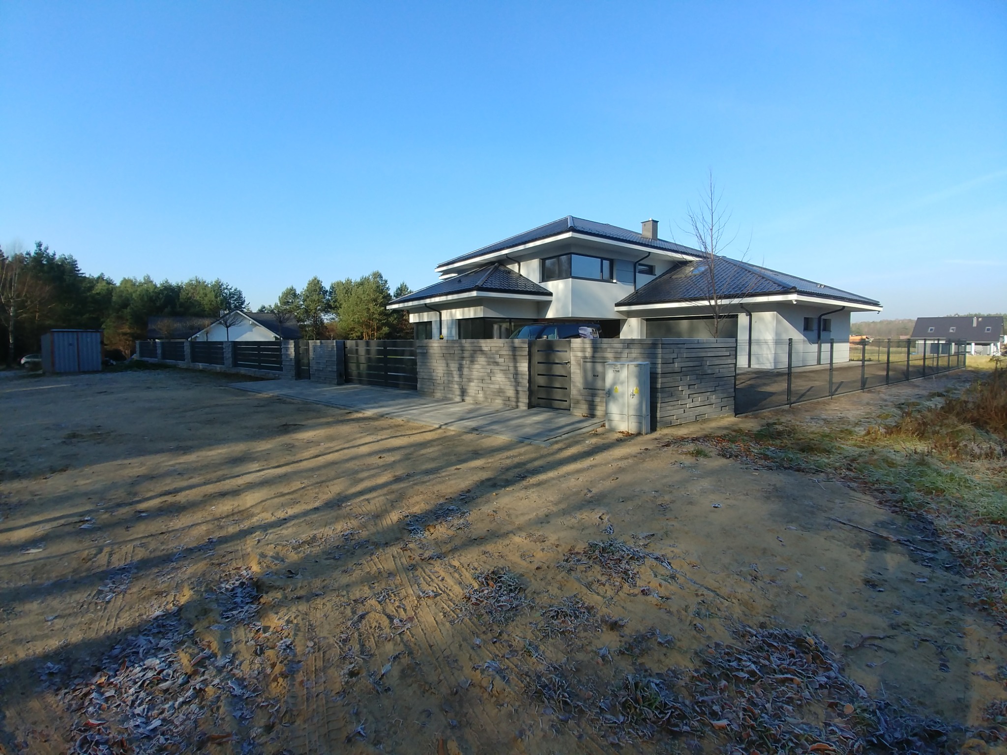 Nowoczesny dom jednorodzinny z szarym ogrodzeniem z betonowych bloczków i metalową siatką, widok z zewnątrz w słoneczny, zimowy poranek, ziemia pokryta szronem.