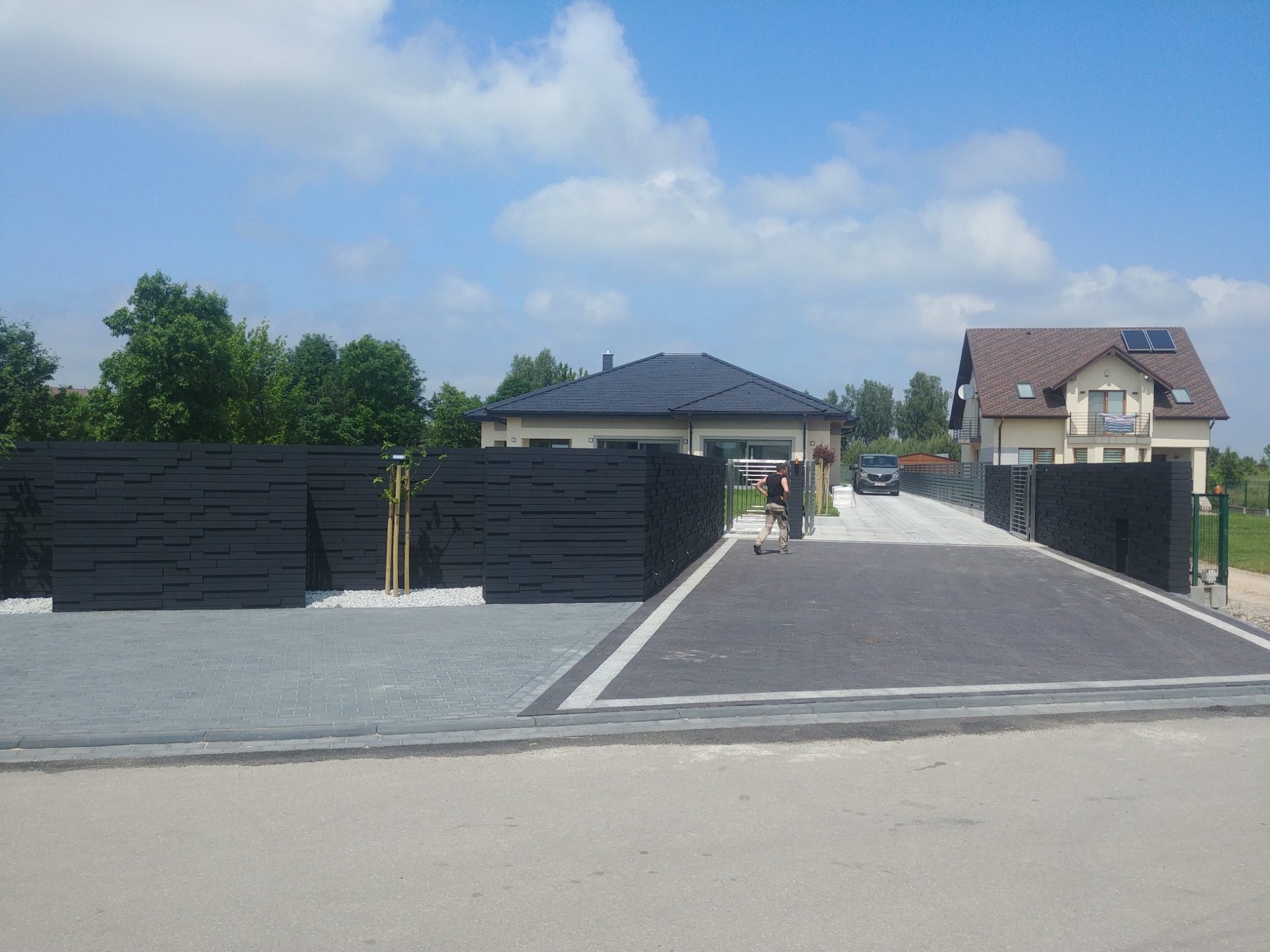 Nowoczesne, czarne ogrodzenie panelowe z poziomych desek, szara kostka brukowa na podjeździe, domy jednorodzinne w tle, słoneczny dzień.