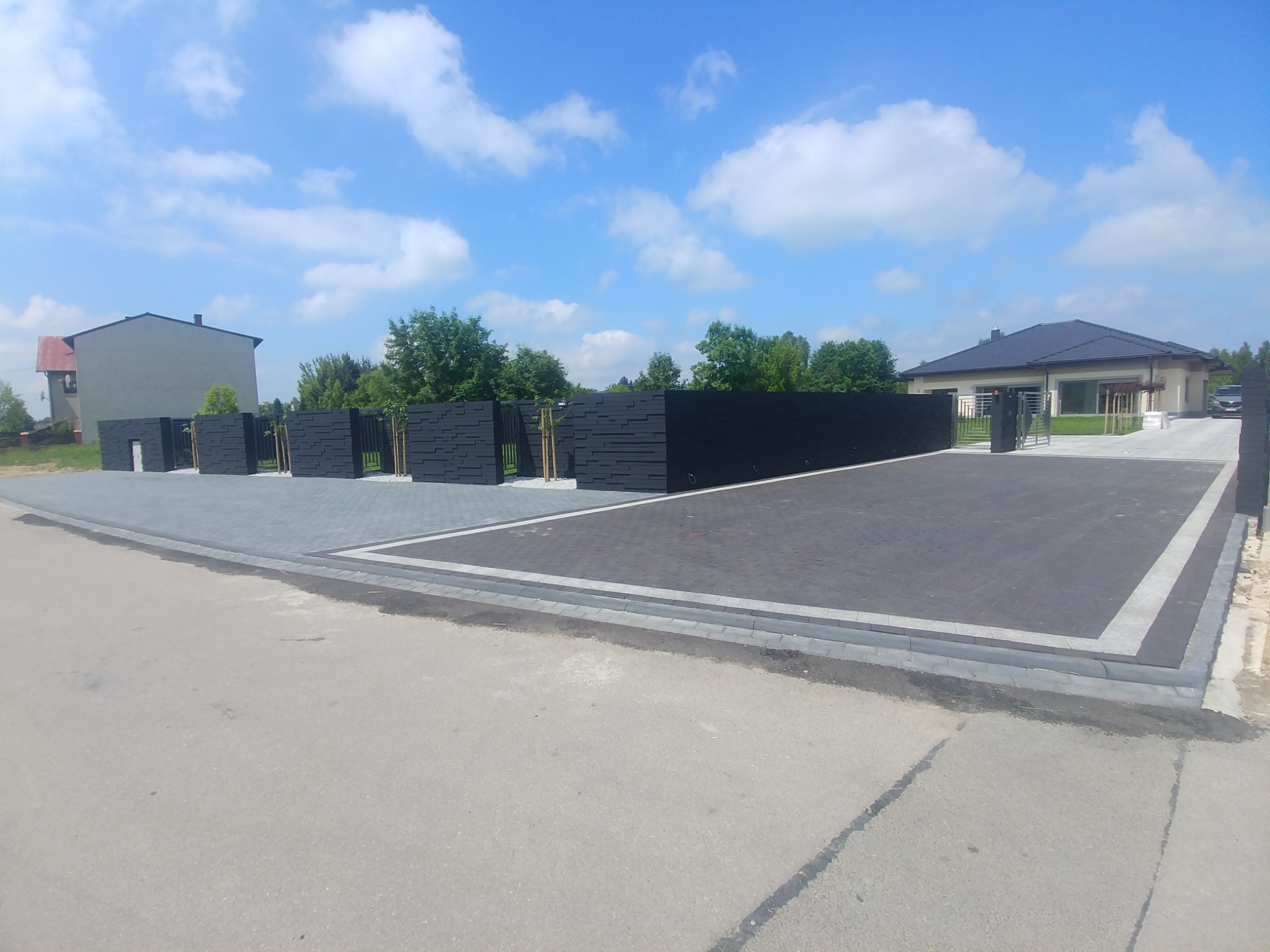 Nowoczesne, czarne ogrodzenie panelowe z poziomych desek, oddzielające posesję z domem jednorodzinnym od ulicy; brama wjazdowa z szarych paneli. Kostka brukowa w dwóch odcieniach szarości.