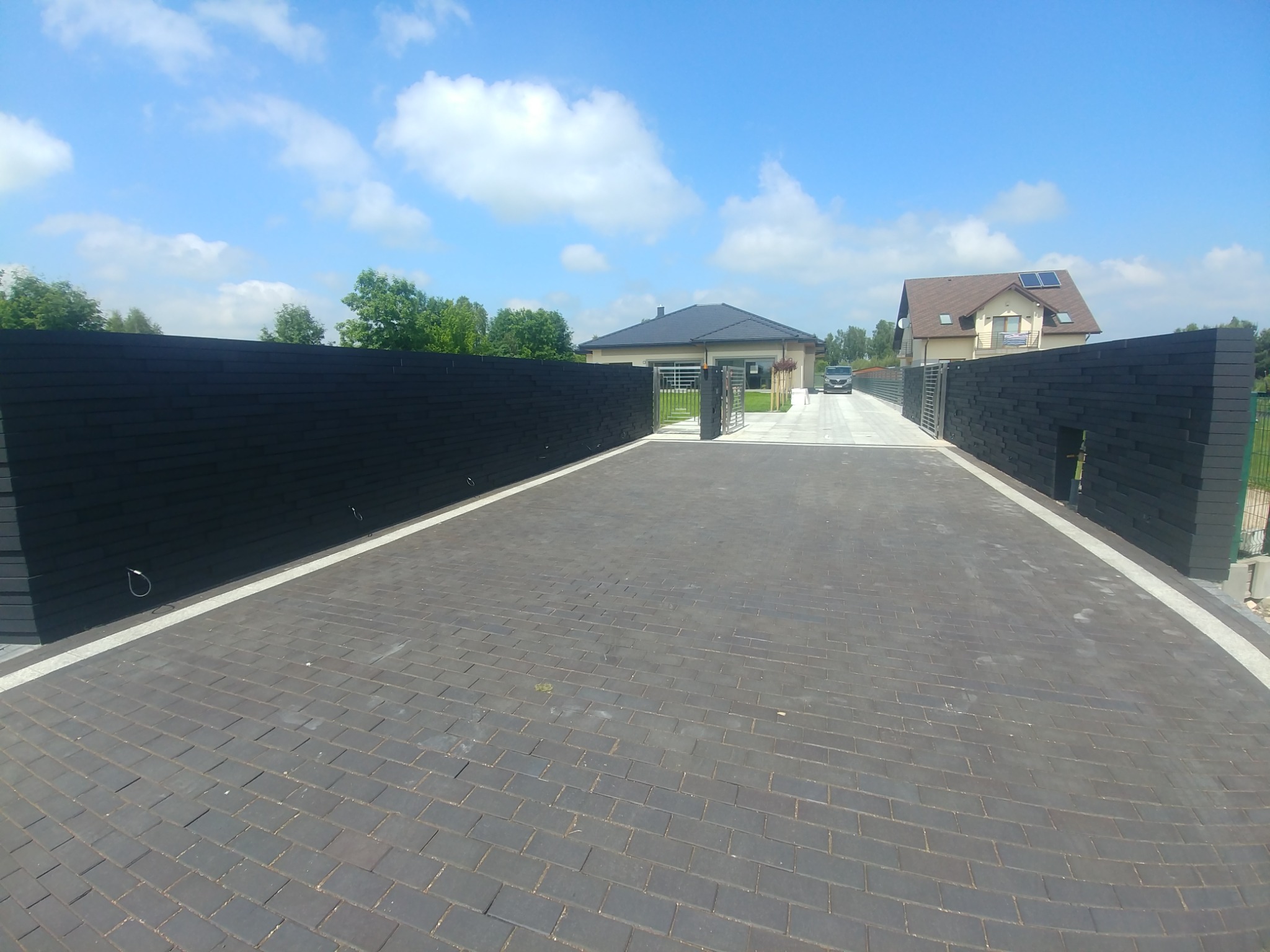 Nowoczesna posesja z czarnym ogrodzeniem z desek kompozytowych, automatyczną bramą wjazdową i brukowanym podjazdem w jodełkę.