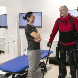 Mężczyzna w czerwonej bluzie, wspomagany egzoszkieletem i laskami, uśmiecha się do kobiety w szarym stroju w jasnym gabinecie z niebieską kozetką i monitorami
