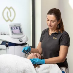 Kobieta w niebieskich rękawiczkach przeprowadza zabieg na twarzy pacjentki leżącej na białym ręczniku, z użyciem specjalistycznego urządzenia INDIVA, w tle widoczne logo Harmony Clinic i drabinka...