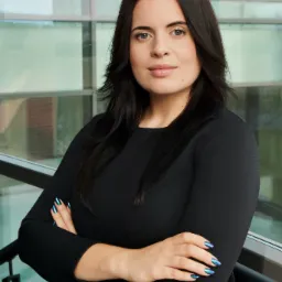 Portret kobiety w czarnej sukience z założonymi rękami na tle przeszklonej ściany.