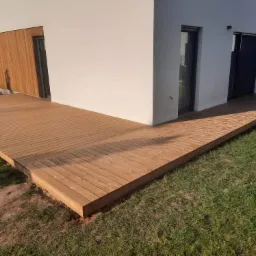 Nowoczesny taras z jasnego drewna przylegający do białego budynku z drewnianą elewacją, widok z poziomu trawnika.