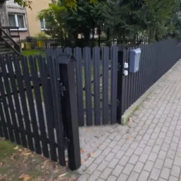 Czarne ogrodzenie z pionowych sztachet i furtką, z zamontowaną szarą skrzynką na listy, widoczne z poziomu chodnika z kostki brukowej, z domem i zielenią w tle.