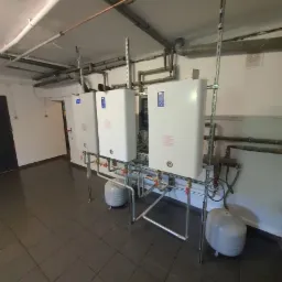 Trzy białe, wiszące kotły grzewcze z podłączonymi rurami i zbiornikami wyrównawczymi, zamontowane w pomieszczeniu z szarą podłogą i białymi ścianami.