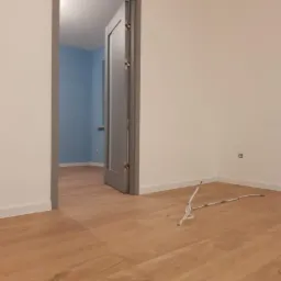 Wnętrze pokoju po remoncie z drewnianą podłogą i szarą ościeżnicą drzwiową, widoczne wystające przewody elektryczne.