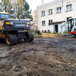 Żółto-czarna koparka Volvo i biało-czerwona koparka Takeushi na placu budowy przed budynkiem z białymi oknami, pracownik w żółtej kamizelce w tle.