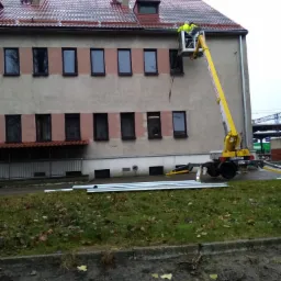 Pracownicy na żółtym podnośniku koszowym przy budynku z czerwonym dachem, prawdopodobnie podczas prac elewacyjnych lub dekarskich, z widocznymi elementami konstrukcyjnymi na ziemi.