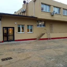 Dwukondygnacyjny budynek o beżowej elewacji z czerwonym pasem u dołu, widoczne okna, klimatyzatory na ścianie, schody zewnętrzne z barierką oraz drabina oparta o ścianę, teren wokół budynku...