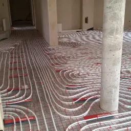 Rozłożone rury ogrzewania podłogowego na siatce z czerwonymi uchwytami, widok z góry na surowy stan wnętrza z betonowym słupem.