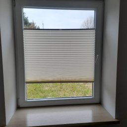 IJM OKNA - Zamknięta, plisowana, biała osłona okienna na oknie PCV, widok na zewnątrz na trawnik i drzewa.