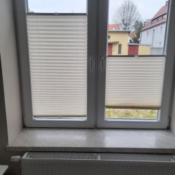 IJM OKNA - Dwuskrzydłowe okno z białymi ramami, z zamontowanymi plisowanymi roletami wewnętrznymi, częściowo zasłoniętymi, widok na zabudowania za oknem.