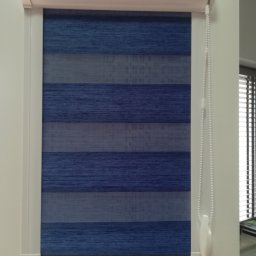 IJM OKNA - Niebiesko-szara roleta okienna typu dzień i noc zamontowana w białej ramie okiennej, z widocznym mechanizmem łańcuszkowym po prawej stronie.